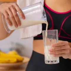 Kobieta w sportowym topie nalewa odżywkę bananową do szklanki. Różne rodzaje odżywek to klucz do regeneracji po treningu.