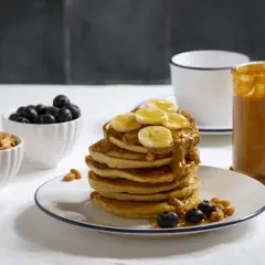 Pancakes z bananem i masłem orzechowym, idealne przepisy z białkiem w proszku. Obok miseczki z borówkami i orzeszkami.