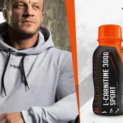 Mężczyzna w szarej bluzie z kapturem obok butelki L-Carnitine 3000 Extreme Shot o smaku gruszka-jabłko.
