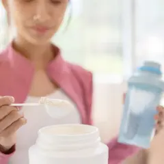 Kobieta przygotowuje odżywkę białkową z mlekiem. Na łyżce jest proszek, obok stoi słoik i shaker.