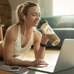 Uśmiechnięta kobieta w słuchawkach pije odżywkę białkową, szukając w internecie odpowiedzi na pytanie: czy białko można pić na noc.