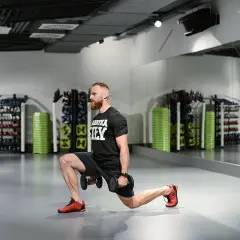 Mężczyzna wykonuje wykroki z hantlami w siłowni. W tle stojaki z obciążeniem i lustra.