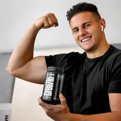 Uśmiechnięty młody mężczyzna pokazuje biceps, trzymając w dłoni opakowanie kreatyny. Idealne wsparcie dla nastolatków budujących masę mięśniową.