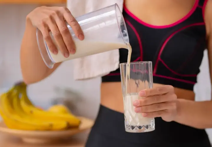 Kobieta w sportowym topie nalewa odżywkę bananową do szklanki. Różne rodzaje odżywek to klucz do regeneracji po treningu.