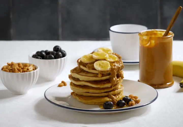 Pancakes z bananem i masłem orzechowym, idealne przepisy z białkiem w proszku. Obok miseczki z borówkami i orzeszkami.