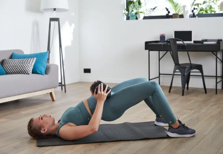 Kobieta wykonuje glute bridge ćwiczenie z obciążeniem na biodrach, leżąc na macie w domowym zaciszu.