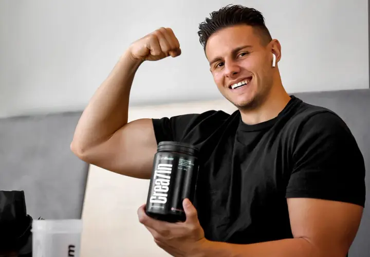 Uśmiechnięty młody mężczyzna pokazuje biceps, trzymając w dłoni opakowanie kreatyny. Idealne wsparcie dla nastolatków budujących masę mięśniową.