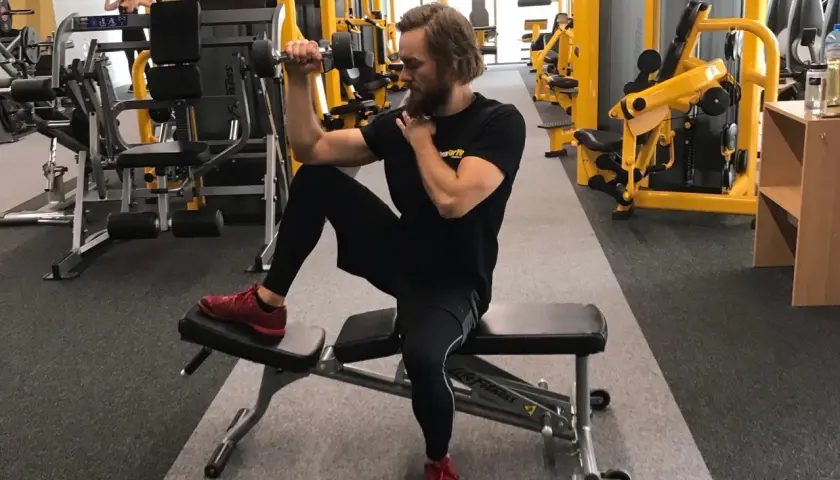 Mężczyzna ćwiczy biceps z hantlem, siedząc na ławce. Dba o balans strukturalny, angażując całe ciało.