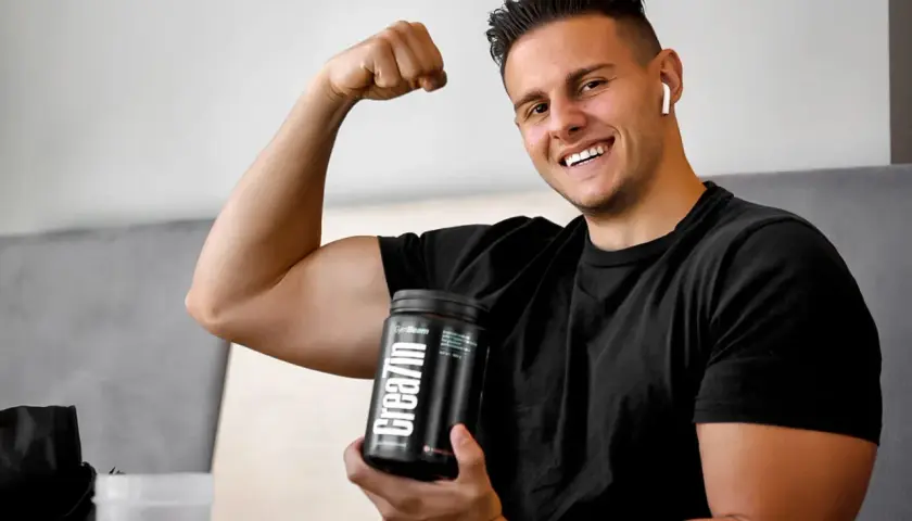 Uśmiechnięty młody mężczyzna pokazuje biceps, trzymając w dłoni opakowanie kreatyny. Idealne wsparcie dla nastolatków budujących masę mięśniową.