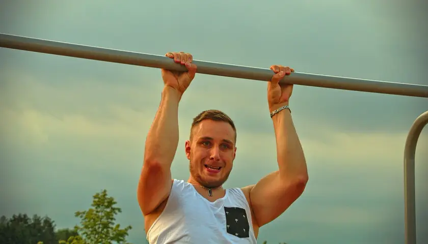 Mężczyzna z uśmiechem wykonuje podciąganie na drążku. To jego plan na dziś, by wzmocnić mięśnie.
