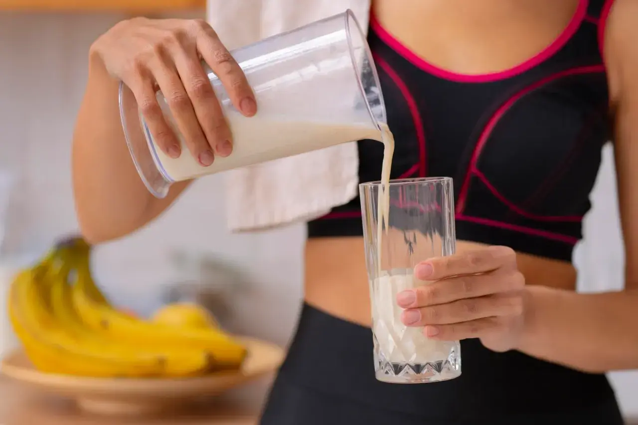 Kobieta w sportowym topie nalewa odżywkę bananową do szklanki. Różne rodzaje odżywek to klucz do regeneracji po treningu.