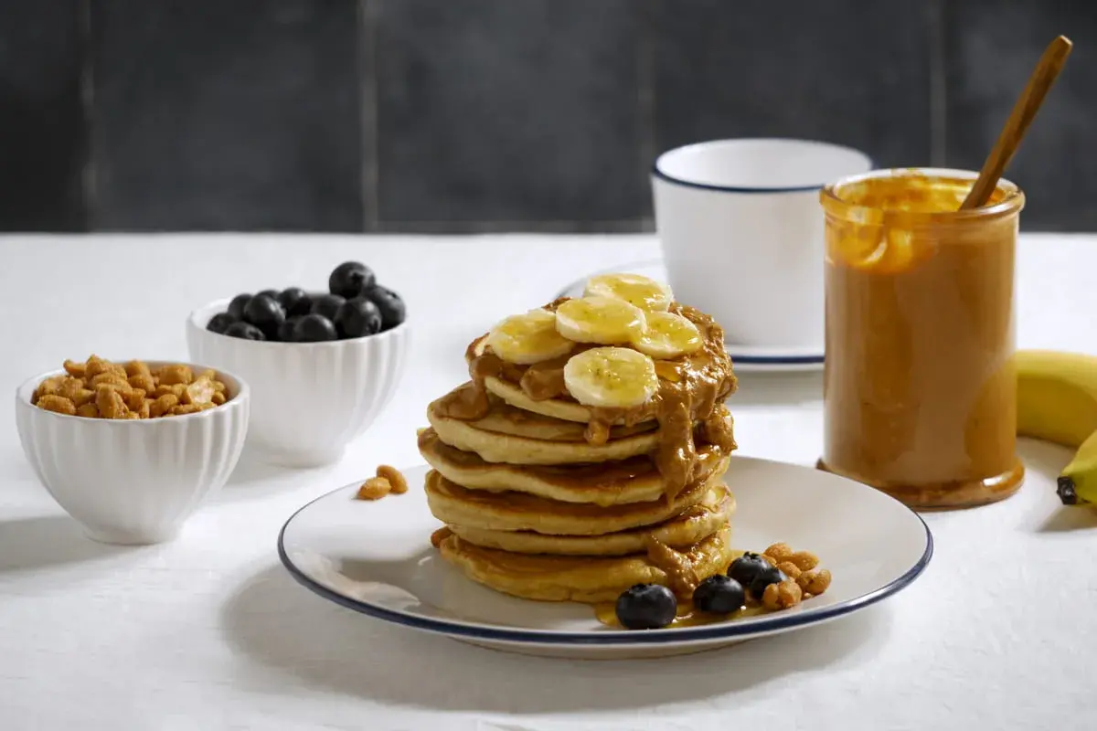 Pancakes z bananem i masłem orzechowym, idealne przepisy z białkiem w proszku. Obok miseczki z borówkami i orzeszkami.