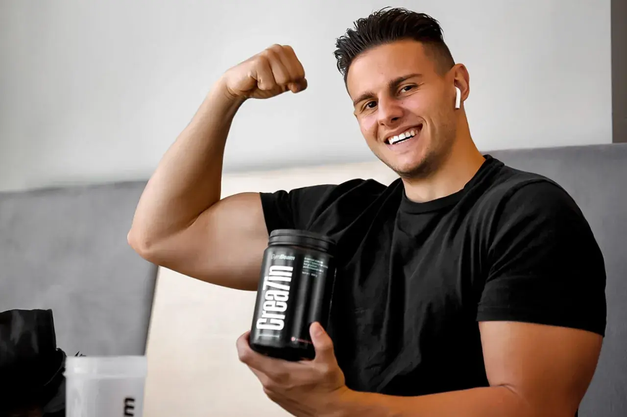Uśmiechnięty młody mężczyzna pokazuje biceps, trzymając w dłoni opakowanie kreatyny. Idealne wsparcie dla nastolatków budujących masę mięśniową.