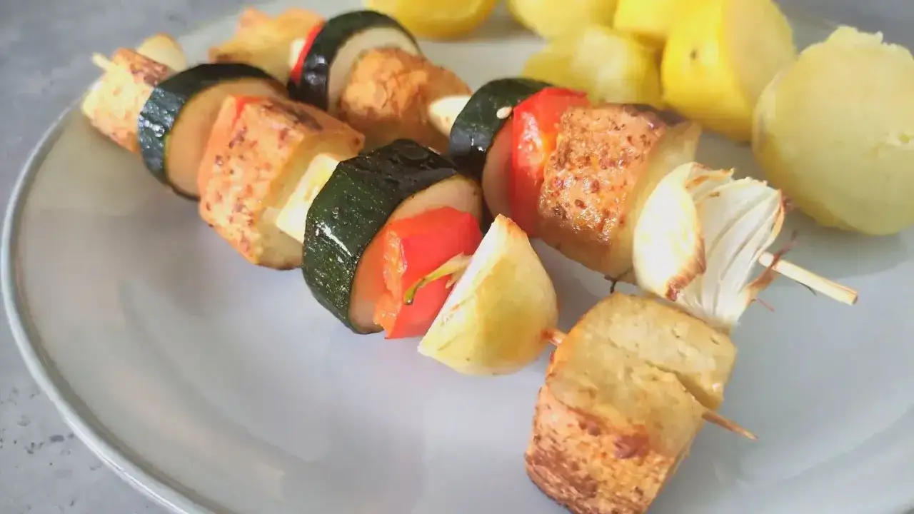 Szaszłyki z tofu, cukinii, papryki i cebuli na talerzu, obok gotowane ziemniaki.